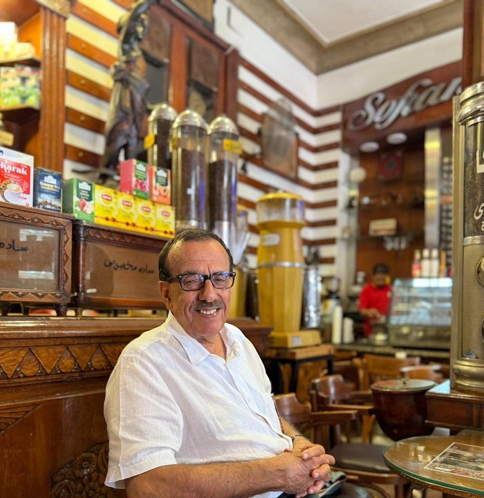 WhatsApp Image 2026-07-12 at 125256 PM - عروبة الاخباري | Oroba News Smiling man with glasses sits in a wooden café or shop, counter and beverage dispensers behind him.