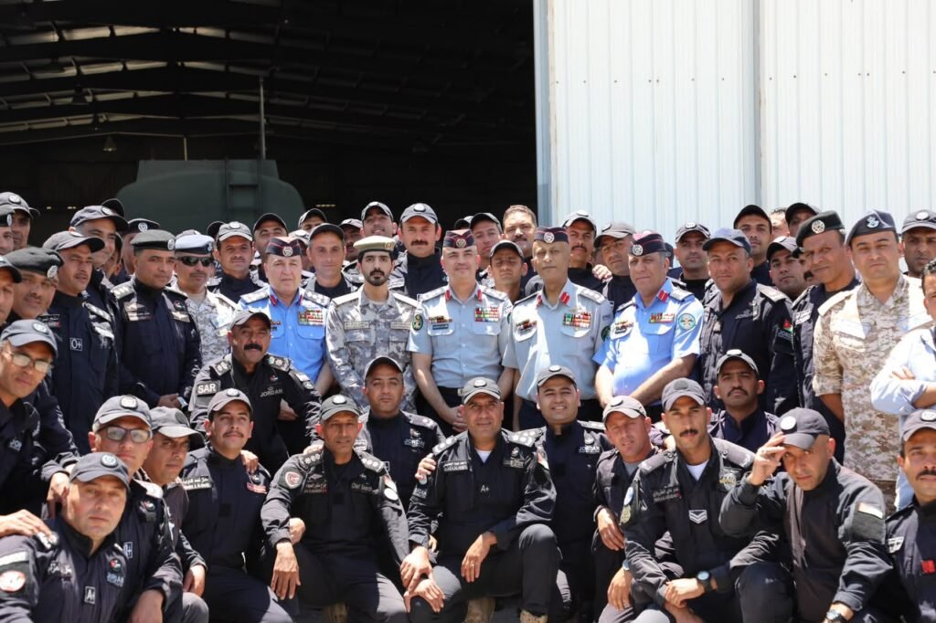Group photo of diverse military personnel in uniform posing outside a hangar, many wearing caps and patches, with some in black, blue, and desert camouflage outfits.