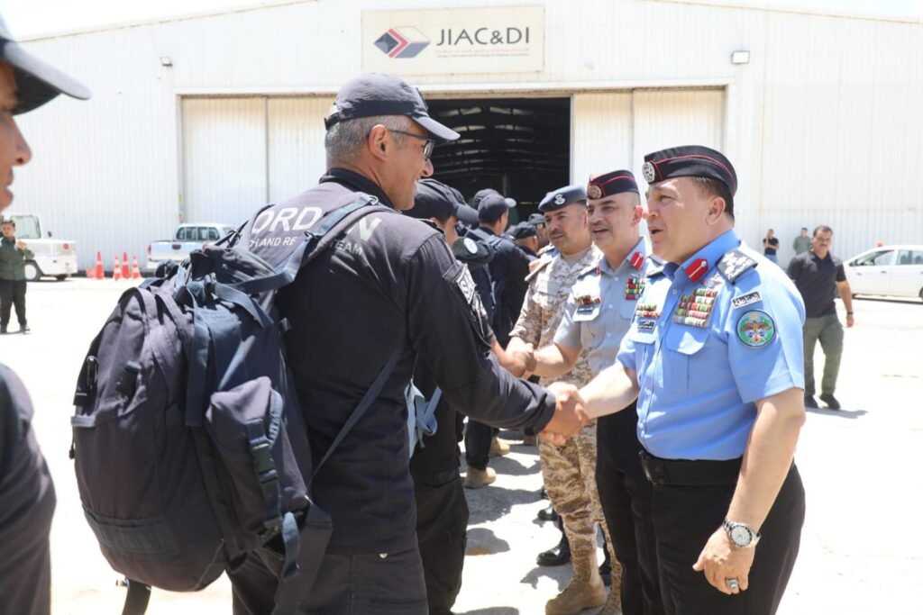 Uniformed security forces shake hands in front of a large hangar, sign visible above reading JIAC&DI, suggesting a briefing or ceremony.