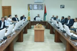 WhatsApp Image 2026-07-14 at 110744 AM - عروبة الاخباري | Oroba News Group of professionals around a U-shaped conference table with laptops, microphones, and a central speaker; flags and portraits on the wall behind.
