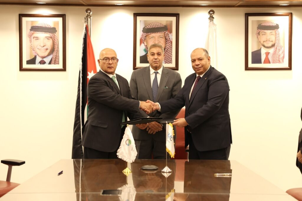 Two men in dark suits shake hands over a folder during a formal signing, while a third man looks on in a conference room with flags and framed portraits behind them.