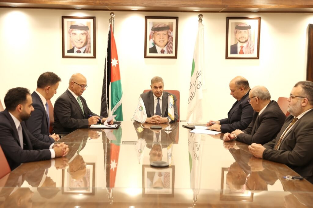 Eight men in suits sit around a polished glass conference table for a formal meeting, with the Jordan flag behind the center chair.