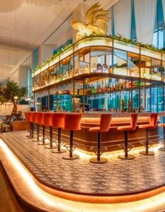 A modern hotel bar with a curved illuminated counter and red stools arranged along the edge, glass shelves stocked with bottles, and a large golden eagle statue atop the display.