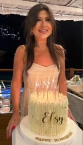 WhatsApp Image 2026-07-15 at 123050 AM1 - عروبة الاخباري | Oroba News Smiling woman in a light beige dress stands behind a white, iced birthday cake with candles and the name 'Elsy' written on it.