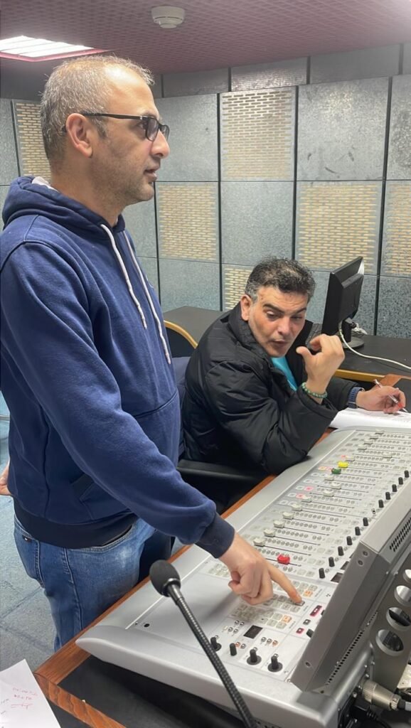 WhatsApp Image 2026-07-15 at 80515 PM2 - عروبة الاخباري | Oroba News Two men in a radio studio: one in a blue hoodie stands and points at a large mixing console while the other sits, talks on a phone, and writes notes.
