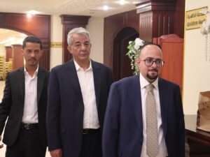Three men in suits walk through a formal hall; center man has gray hair, flanked by two others in dark jackets.