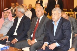 Three men in suits seated at a formal event, with one man wearing a traditional red-and-white headscarf on the left and others in business attire in the foreground.