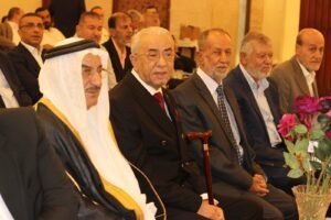 Group of men in suits seated at a formal event, including a man in traditional Arab attire with a wooden cane in front of him and pink flowers nearby.