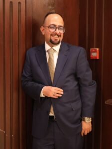 Man in a navy suit and patterned tie smiling, standing against wooden panels with hands in front.