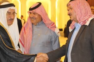 Three men in traditional Arab dress shake hands and smile in a warmly lit indoor setting.