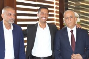 Three men in suits posing together at a formal event; center man smiles as the other two stand beside him.