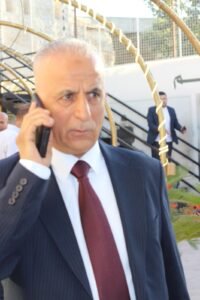 Man in a dark pinstripe suit and burgundy tie talks on a cellphone outdoors, with a decorative circular frame in the background.