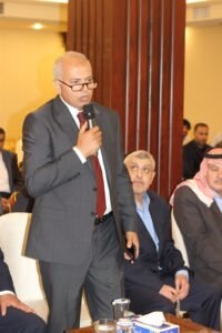 Man in a gray suit and red tie speaks into a handheld microphone at a formal event, with seated attendees in the background.