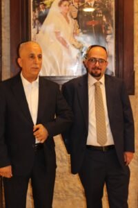 Two men in dark suits stand side by side in a formal setting, with a large framed bridal portrait behind them on the wall.