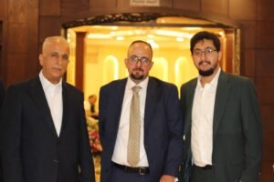 Three men in suits posing for a photo in a warmly lit lobby or entrance area, smiling at the camera.