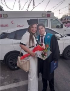 WhatsApp Image 2026-07-16 at 113730 AM - عروبة الاخباري | Oroba News Graduate in cap and gown poses with a bouquet-wielding woman beside a white car and an ambulance in the background.