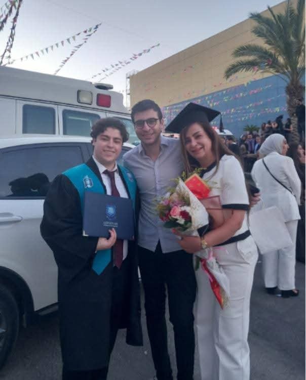 WhatsApp Image 2026-07-16 at 113730 AM1 - عروبة الاخباري | Oroba News Three people pose at a graduation ceremony: a graduate in cap and gown holding a diploma, flanked by a man and a graduate in white holding flowers.