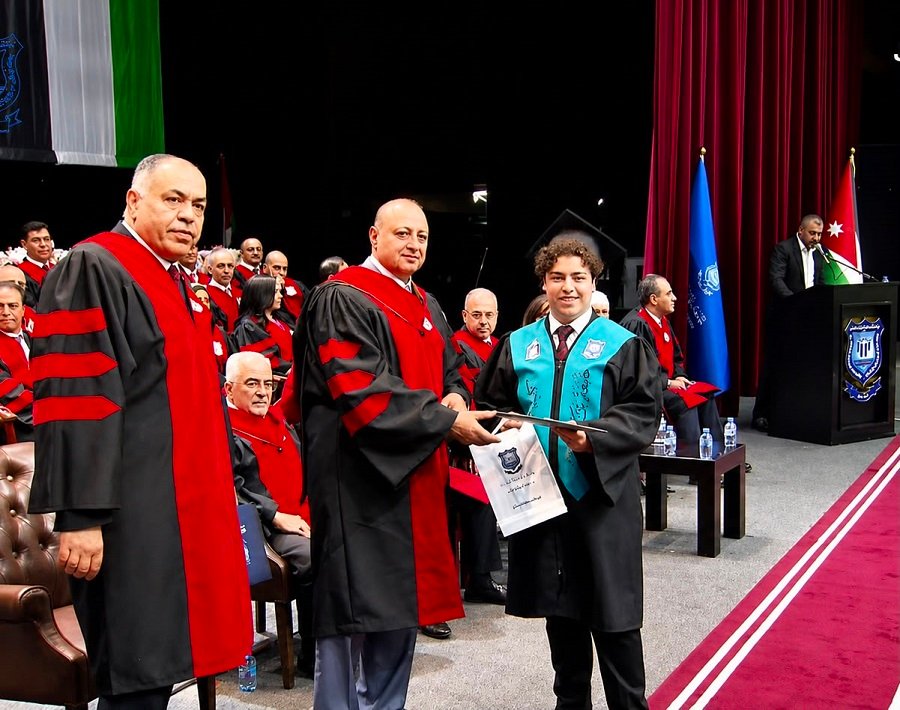 WhatsApp Image 2026-07-16 at 23001 PM - عروبة الاخباري | Oroba News Graduate in a black gown with teal stole receives a diploma from a faculty member in red robes during a graduation ceremony on stage.