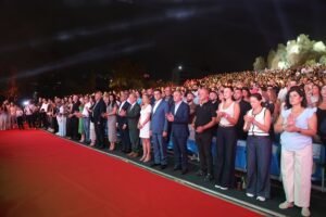 WhatsApp Image 2026-07-17 at 121755 PM1 - عروبة الاخباري | Oroba News Nighttime outdoor ceremony with a large crowd standing and clapping along a red carpeted area outside a stadium or arena.Spotlights illuminate the audience on the hillside above.