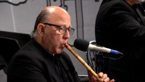 Man in a black suit plays a wooden recorder into a microphone during a performance.