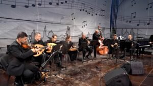 Stage view of a string ensemble performing in black attire, with violins, guitars/oud, a cello and a double bass seated in a row on a concert stage.