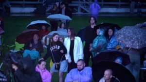 Crowd of people under umbrellas at a rain-soaked outdoor event on a grassy field at night; some sit in chairs.