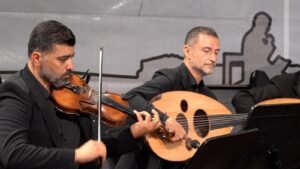 Two male musicians in black suits perform on stage: one playing a violin, the other an oud, with music stands nearby.