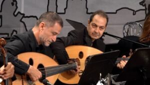 Two male musicians in black, playing oud guitars and reading sheet music during a performance.
