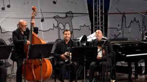 Jazz trio on stage: bassist standing left, two seated players with percussion and violin, grand piano on right, backdrop with city skyline and musical notes