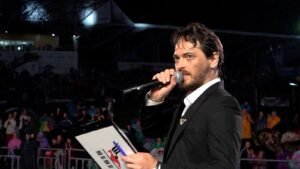 Male singer in a black suit performing on stage at night, holding a microphone and lyric sheet before a crowd.
