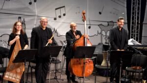 Four musicians on stage pose with their instruments: hammered dulcimer player, flute player, double bass player, and pianist, all smiling in black attire.