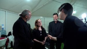 Interviewer hands a microphone to a smiling woman as three men in black suits watch backstage in a tented area.