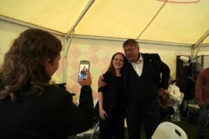 Smiling woman and man pose for a photo inside a yellow tent; a person in the foreground takes their picture with a smartphone.