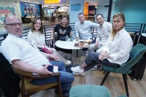 Group of six adults seated around a circular table in a mall café, chatting with drinks and snacks on the table, Dunkin' sign in background.