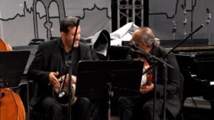 Two male musicians in black suits share a laugh on stage, one holding a tambourine and the other a violin while facing each other near a music stand and microphones.