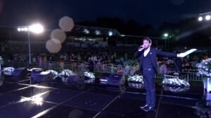 Male performer in a dark suit singing into a microphone on a rain-soaked outdoor stage, with a cheering audience in a stadium behind him and bright stage lights above.