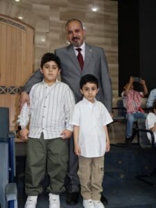 WhatsApp Image 2026-07-18 at 104002 AM 3 - عروبة الاخباري | Oroba News Man in a gray suit stands behind two boys, arms around them, posing for a photo on an indoor stage. Audience members are in the background.