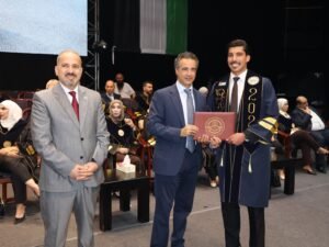 WhatsApp Image 2026-07-18 at 104002 AM - عروبة الاخباري | Oroba News Graduate in a navy gown with gold trim receives a diploma from a man in a blue suit on stage during a ceremony; audience seated in background.