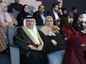 WhatsApp Image 2026-07-18 at 104002 AM 4 - عروبة الاخباري | Oroba News Man in traditional Arab dress (black bisht with gold trim and white ghutra) sits beside a smiling woman in a beige hijab, in a theater seating area.