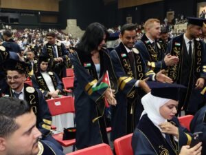 WhatsApp Image 2026-07-18 at 104003 AM 1 - عروبة الاخباري | Oroba News Graduates in navy gowns with gold trim stand and sit in a large arena, some waving Jordan flags as a crowd watches a graduation ceremony.