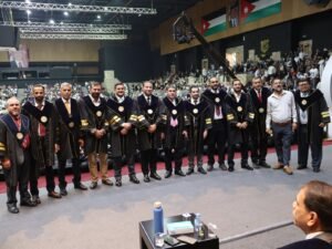 WhatsApp Image 2026-07-18 at 104003 AM 3 - عروبة الاخباري | Oroba News A row of graduates in black gowns with gold trim poses on a stage in a large indoor graduation ceremony hall.