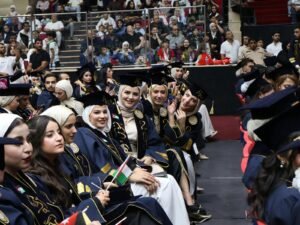 WhatsApp Image 2026-07-18 at 104003 AM 4 - عروبة الاخباري | Oroba News Graduates in blue gowns and caps seated in a ceremony hall, some smiling and waving, with audience behind glass barriers in the background and small flags in hand.
