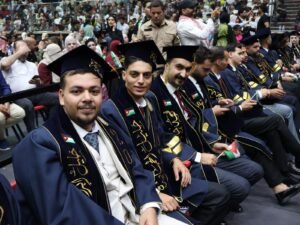 WhatsApp Image 2026-07-18 at 104003 AM 6 - عروبة الاخباري | Oroba News Front-row graduates in navy gowns and mortarboards with gold embroidery, holding small Jordan flags at a crowded ceremony.