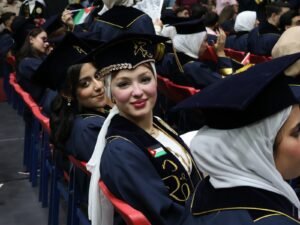 WhatsApp Image 2026-07-18 at 104003 AM 7 - عروبة الاخباري | Oroba News Two graduates in navy gowns and caps sit in a crowded ceremony, smiling at the camera; the woman in front wears a white headscarf or veil.