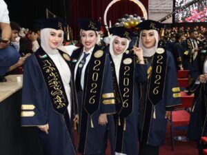 WhatsApp Image 2026-07-18 at 104004 AM 1 - عروبة الاخباري | Oroba News Four graduates in navy gowns and caps pose at a graduation ceremony, with a stage and balloons behind them.