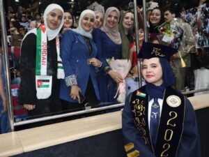 WhatsApp Image 2026-07-18 at 104004 AM 2 - عروبة الاخباري | Oroba News Graduate in a dark blue gown and cap with gold embroidery, smiling at the camera in front of a cheering crowd; Jordan flag scarf draped over a woman beside her.