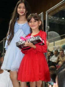 WhatsApp Image 2026-07-18 at 104004 AM - عروبة الاخباري | Oroba News Two smiling girls in dresses stand on a stage; the girl in red holds a boxed Tom Ford gift with a pink ribbon.