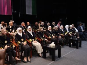 WhatsApp Image 2026-07-18 at 104004 AM 3 - عروبة الاخباري | Oroba News Group of graduates in black gowns with gold trim, many wearing white hijabs, seated in an auditorium for a ceremony.