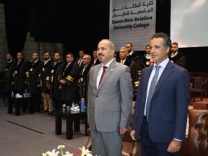 WhatsApp Image 2026-07-18 at 104004 AM 5 - عروبة الاخباري | Oroba News Two men in suits stand in the foreground at a formal graduation ceremony while a line of graduates in black gowns with gold trim stand behind them on stage.