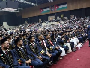 WhatsApp Image 2026-07-18 at 104004 AM 6 - عروبة الاخباري | Oroba News Rows of graduates in navy blue gowns and caps with gold embroidery sit on a red-carpeted stage at a large indoor ceremony, audience in the stands behind them.